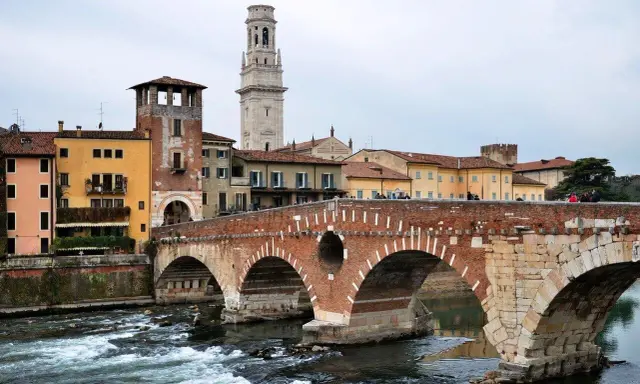 En vacker tegelbro korsar en flod i Verona, Italien. Färgglada hus och ett högt klocktorn syns i bakgrunden.
