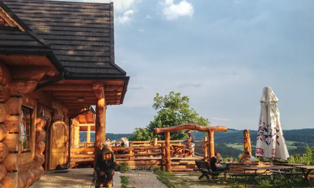 Drewniana bacówka Jamna z tarasem, gdzie goście jedzą, a obok stoi parasol. Menu bacówki Jamna kusi widokiem.