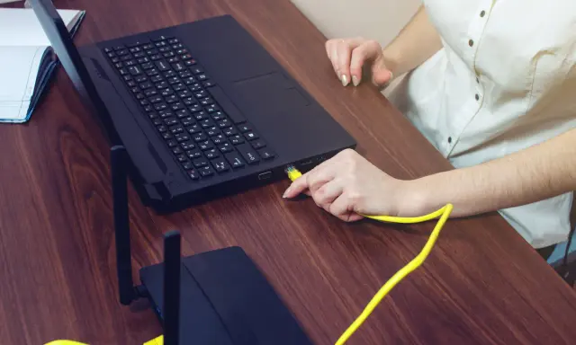 Ręka podłącza żółty kabel do laptopa, pokazując, czy można podłączyć monitor do laptopa. Obok leży router.