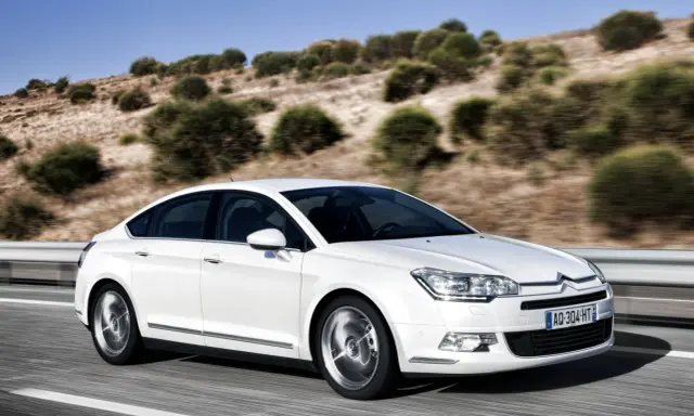 Biały Citroen C5 2.0 HDI pędzi po pustej drodze, otoczony suchymi, pagórkowatymi krajobrazami.
