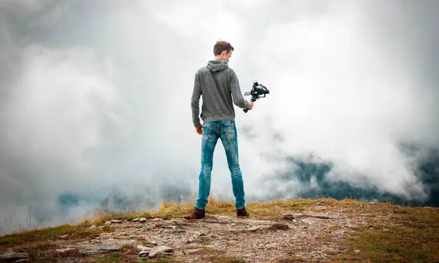 Joven en la cima de una montaña, sosteniendo un **gimbal** para estabilizar su cámara y capturar la vista entre las nubes.