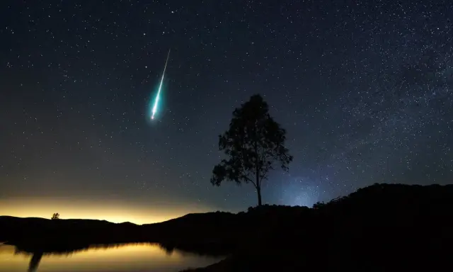 Un **obiettivo per fotografare le stelle** cattura una scia luminosa nel cielo notturno, con un albero solitario in silhouette e un lago calmo.