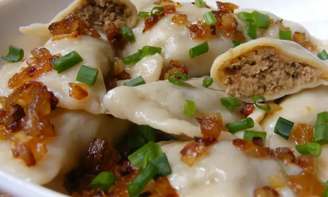 Pyszne pierogi z mięsem, okraszone skwarkami i szczypiorkiem, w białej misce.