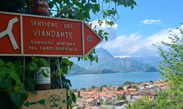 Segnale arancione indica "Sentiero del Viandante" con vista su un lago e montagne.