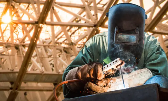 Soldador con máscara y guantes realiza soldadura con electrodo, creando chispas y humo bajo una estructura metálica iluminada por el sol.