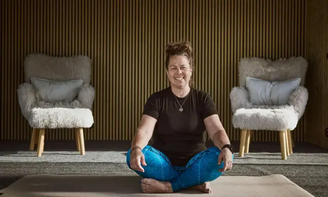 Kvinna i meditationsställning framför en vägg med träribbor. Två fluffiga fåtöljer står på var sin sida. Kanske hon funderar på hur hon ska dekorera hylla i sitt hem.