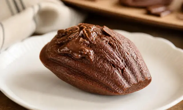 Un biscuit madeleine au chocolat, nappé de glaçage et parsemé de copeaux de chocolat, sur une assiette blanche.