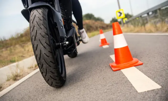 Motociclista practica maniobras entre conos naranjas. La rueda delantera de la moto, con su banda de rodadura, se ve de cerca.