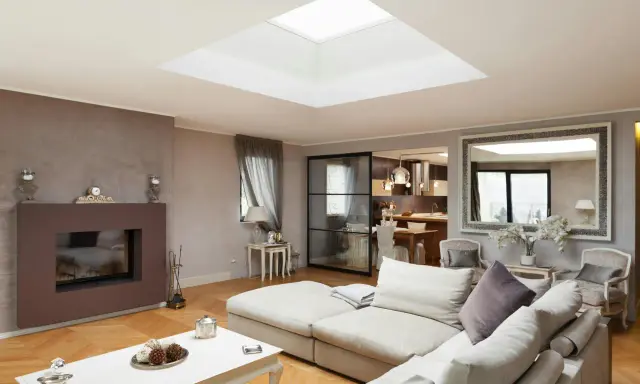 Un salon moderne avec un grand puit de lumière au plafond, un canapé confortable, une cheminée et une cuisine ouverte.