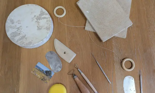 Keramikverktyg och en lerklump på ett träbord. Här finns allt för att visa vad man kan dreja: skärtråd, träformar, metallverktyg och en svamp.