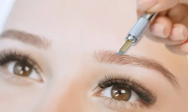 Main d'une esthéticienne réalisant un **tatouage sourcils poil par poil** avec un outil fin. Gros plan sur un œil et un sourcil.