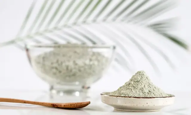 Une cuillère en bois repose à côté d'un petit bol rempli d'argile verte à boire. En arrière-plan, un grand bol contient également cette poudre naturelle.