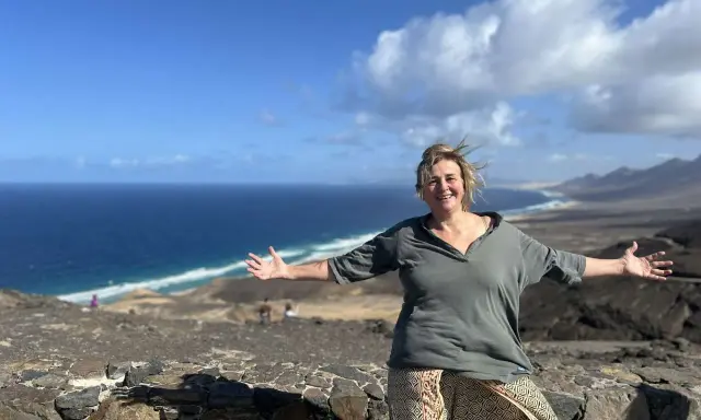 Donna sorridente con le braccia aperte su un paesaggio costiero, un'espressione di gioia che contrasta la solitudine interiore.