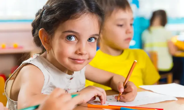 Uśmiechnięta dziewczynka z niebieskimi oczami rysuje kredką. Jej przeniesienie dziecka do innego przedszkola nie zasmuciło jej.