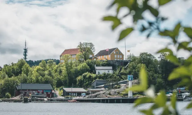 Widok na wybrzeże w Kristiansand, Norwegia. Żółte budynki na wzgórzu, czerwony klub kajakowy i wieża telekomunikacyjna.
