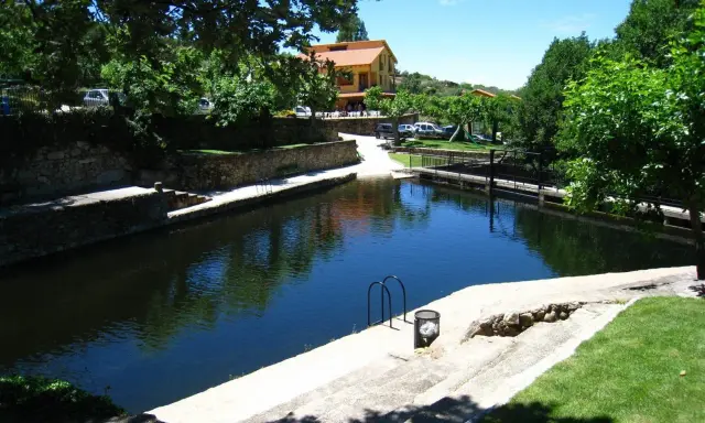 La piscina natural de Casas del Monte, rodeada de vegetación y con un edificio al fondo, invita a un refrescante baño.