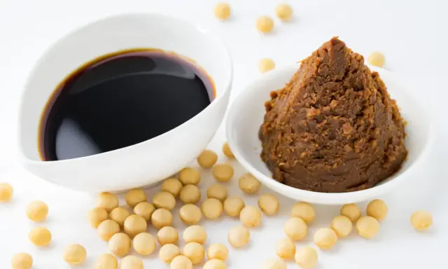 Schüssel mit dunkler Sojasauce und eine mit brauner Miso-Paste, umgeben von Sojabohnen. Eine leckere hefeflocken alternative.