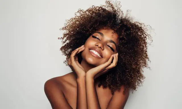 Una mujer sonríe con los ojos cerrados, su cabello rizado enmarca su rostro. Su piel radiante y su expresión serena sugieren cómo parecer más joven.