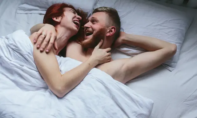 Para kochająca się w łóżku, śmiejąca się. Kobieta z rudymi włosami obejmuje mężczyznę z brodą. Sikanie podczas seksu może być częścią intymnych chwil.
