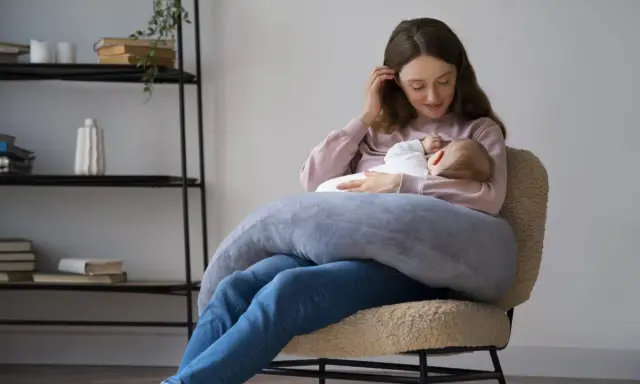 Szukasz idealny fotel do karmienia? Młoda mama z dzieckiem na rękach, otulona poduszką, siedzi na wygodnym, pluszowym fotelu.