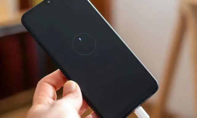 Dłoń trzyma smartfon z ikoną ładowania zwrotnego na czarnym ekranie. Podłączony biały kabel USB.