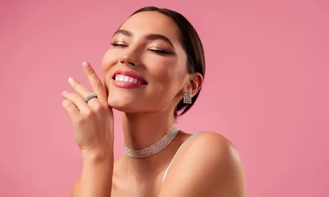 Mujer sonríe con ojos cerrados, luciendo un maquillaje romántico, joyas brillantes y un fondo rosa.