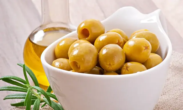 Aceitunas verdes en un cuenco blanco junto a una botella de aceite de oliva y romero. Descubre los aceitunas beneficios y contraindicaciones.