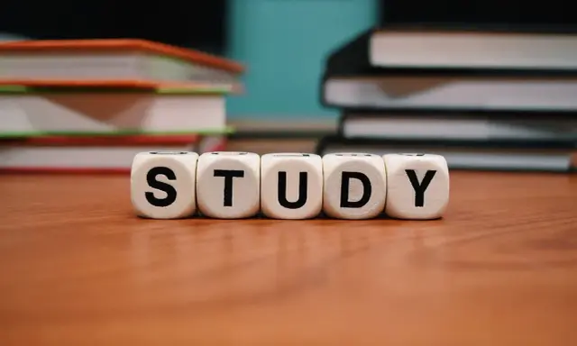 Białe kostki z literami "STUDY" na drewnianym stole, w tle stosy książek.