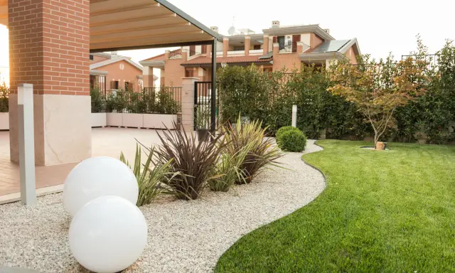 Aiuole davanti casa con ghiaia bianca, sfere luminose e prato verde curato. Sullo sfondo, case moderne e siepi.