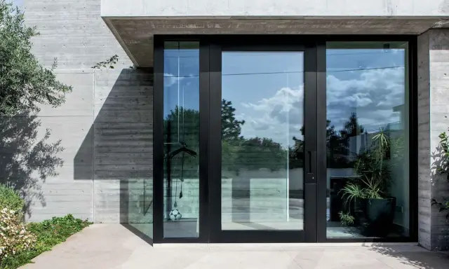 Puerta de entrada moderna con grandes ventanales de cristal, mostrando diferentes tipos de cristales para puertas exteriores que reflejan el cielo y la vegetación.