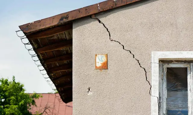 Grieta en pared de edificio, indicando problemas de **seguridad estructural**. Se observa un alero de madera y un cielo azul.