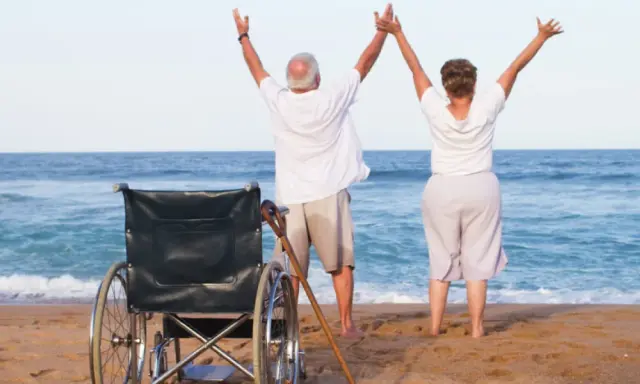Szczęśliwi seniorzy cieszą się urlopem. Wózek inwalidzki i laska na plaży świadczą o tym, że to udany turnus rehabilitacyjny nad morzem.