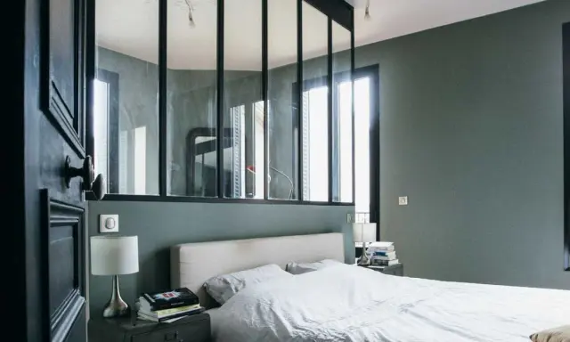 Agencement chambre moderne avec lit blanc, table de chevet industrielle et verrière séparant l'espace.