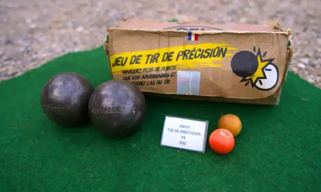 Deux boules de pétanque et un cochonnet sur un tapis vert. La boîte du jeu de tir de précision est visible. Apprenez comment tirer à la pétanque !