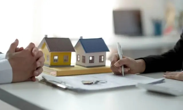 Manos entrelazadas junto a maquetas de casas y un bolígrafo firmando un contrato privado de alquiler.