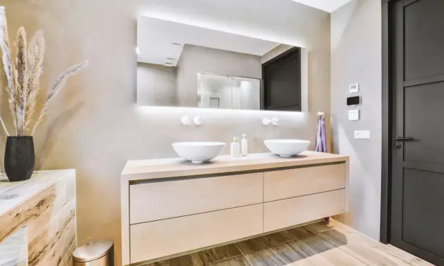 Elegantes baños modernos pequeños con lavabos dobles sobre mueble de madera clara, espejo iluminado y pared texturizada.