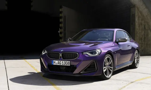 Fioletowe BMW 220i coupe z nowoczesnym designem, zaparkowane na betonowym placu przed ciemnym budynkiem.