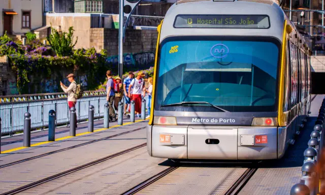 En modern spårvagn från Metro do Porto närmar sig stationen. Passagerare väntar på perrongen, omgivna av grönskande växtlighet.