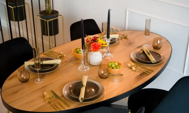 Un dîner élégant est prêt, avec des assiettes sombres, des couverts dorés et des raisins verts. Les art de la table sont mis en valeur par des bougies noires et des fleurs colorées.