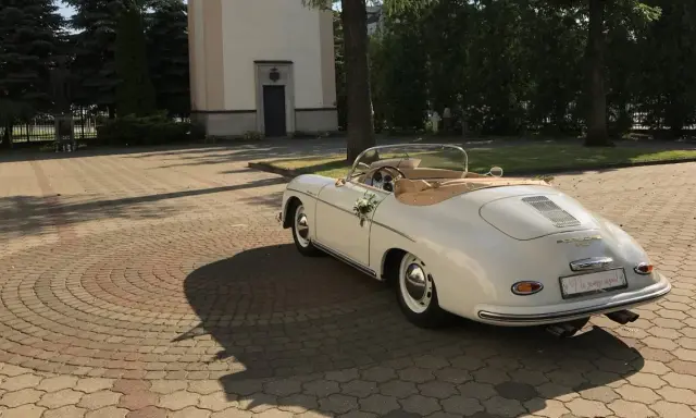 Biały, zabytkowy kabriolet Porsche, udekorowany kwiatami, czeka przed kościołem. To idealny samochod na wesele, gotowy na podróż poślubną.