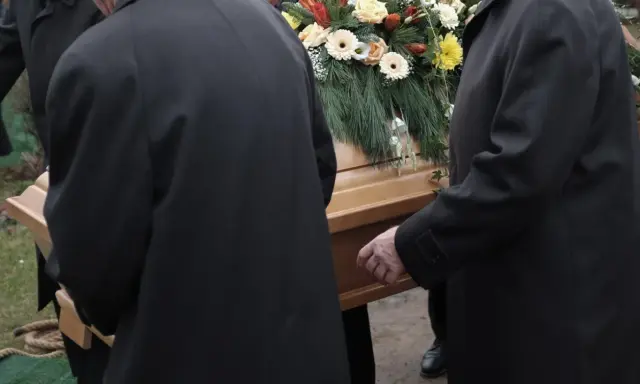 Mężczyźni w czarnych płaszczach niosą trumnę ozdobioną kwiatami. Kondukt pogrzebowy w żałobnej ciszy.