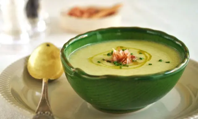 Un refrescante gazpacho de melón, adornado con jamón serrano y cebollino, servido en un cuenco verde con una cuchara dorada.