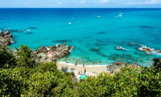 Spiaggia affollata con gente che nuota e prende il sole, barche in mare e vegetazione lussureggiante. Un angolo di paradiso del **calabria mare**.
