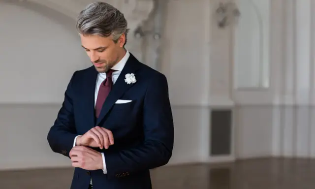 Elegancki mężczyzna w granatowym garniturze i białej koszuli, z bordowym krawatem i butonierką, przygotowuje się na wesele.
