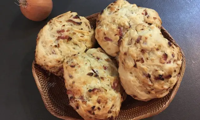 Quatre pains aux lardons et oignons dorés dans un panier en osier, avec un oignon cru à côté.
