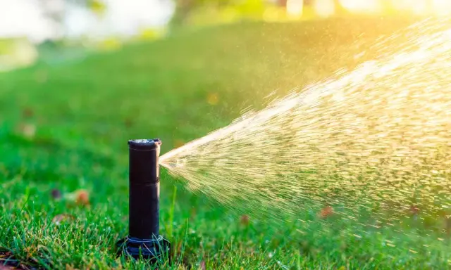 Aspersor de riego automático en acción, rociando agua sobre césped verde bajo la luz del sol.