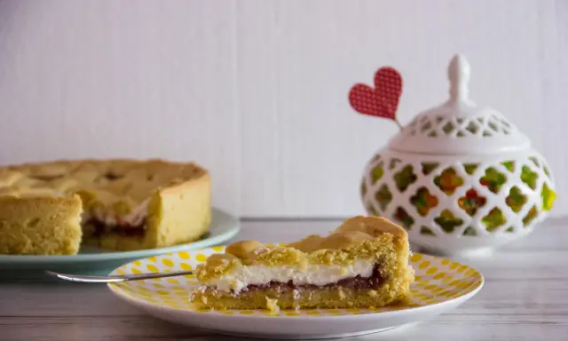 Slice of crostata with ricotta and jam on a yellow polka dot plate, with a whole crostata in the background.