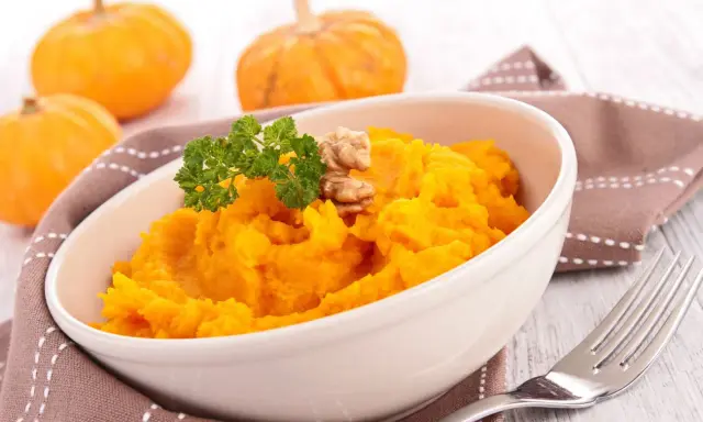 Un tazón de cremoso **puré de calabaza al horno**, adornado con perejil y nueces. Al fondo, calabazas pequeñas y una servilleta marrón.