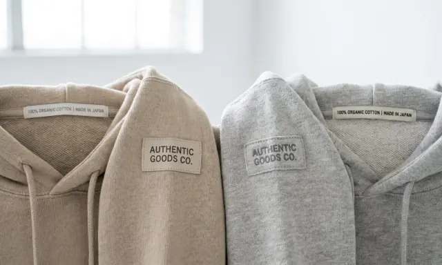 Dwa bluzy z kapturem, beżowa i szara, z naszywką "AUTHENTIC GOODS CO." i metką "100% ORGANIC COTTON | MADE IN JAPAN". Oryginalna metka Champion.