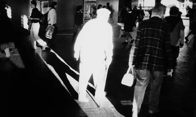 A stark black and white image by Trent Parke captures a blurred figure in a white hat and coat, walking with a cane. Other figures are also blurred, suggesting movement and a fleeting moment.